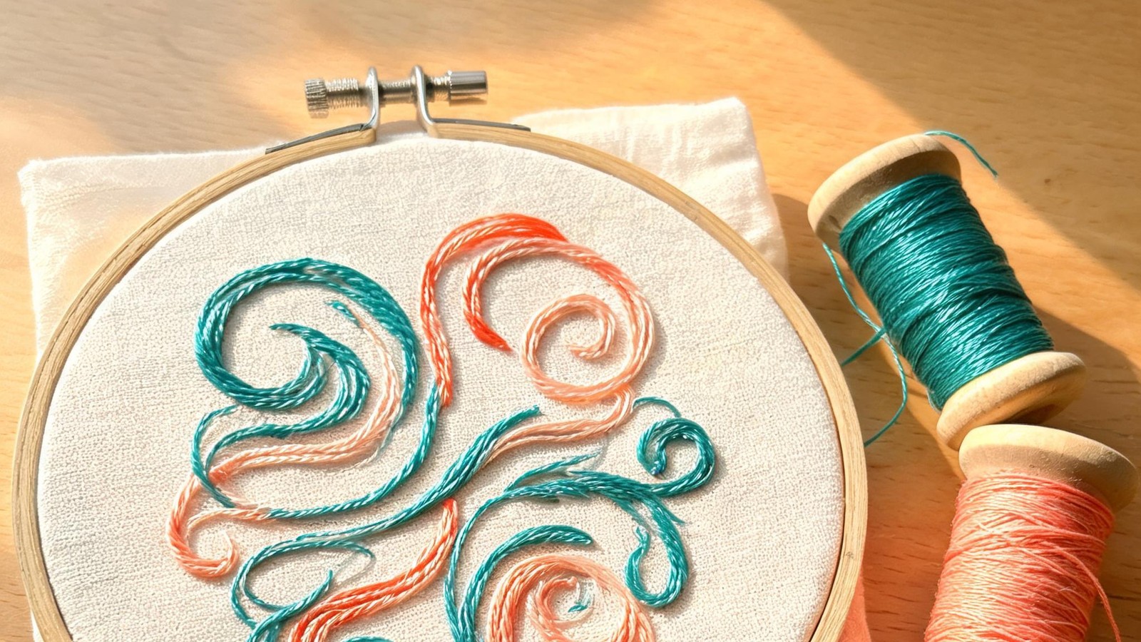Illustration of an embroidery hoop with teal and coral satin-stitch curves and two thread spools on a wooden table.