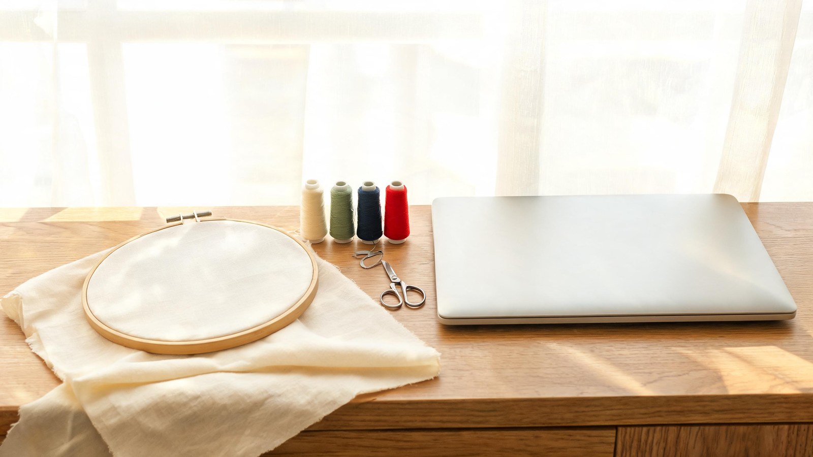 Illustration of an embroidery hoop on plain fabric, thread spools, scissors, and a closed laptop on a wooden table.